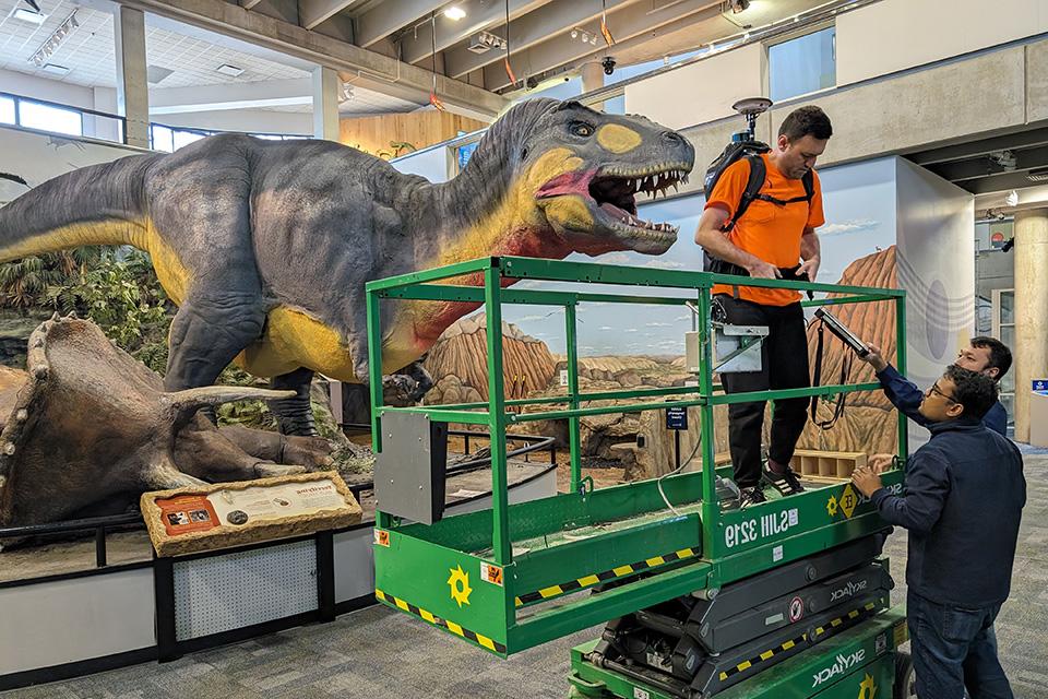 A student st和s on a lift with a large piece of equipment on his back in front of one of the latex dinosaurs.